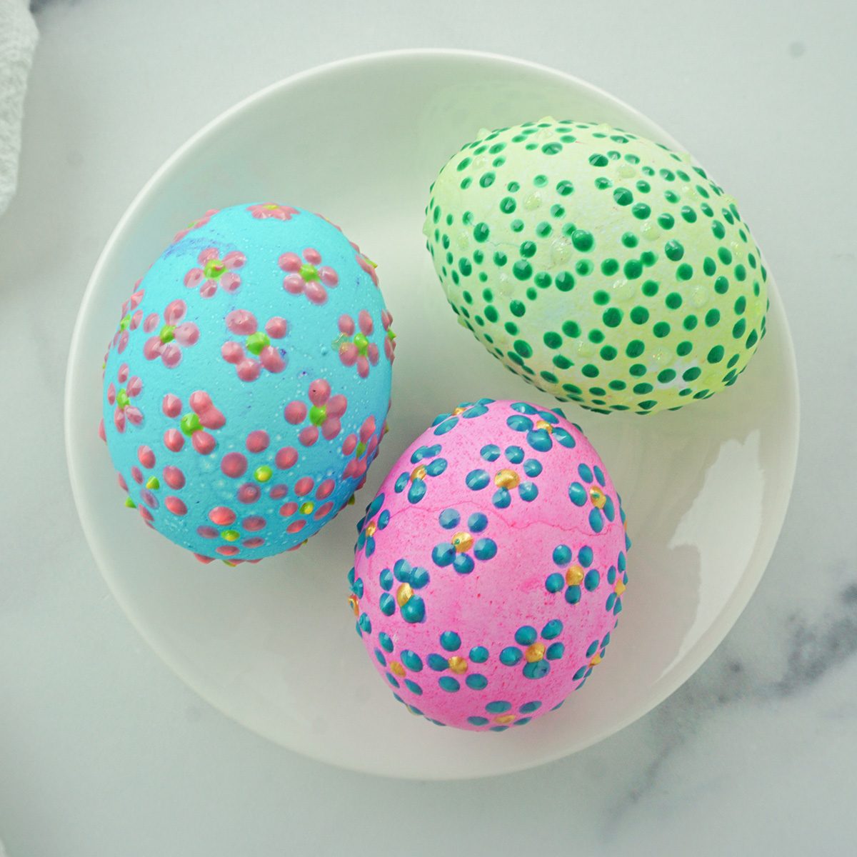 Three decorated eggs on a white plate. One is blue with pink and green floral patterns, another is pink with blue and pink flowers, and the third is green with small, multicolored dots. They are set on a marble surface.