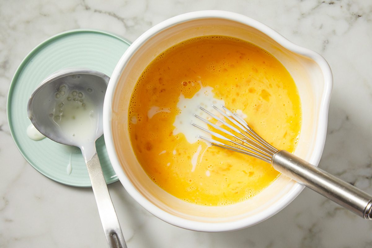 A white bowl with orange liquid and a metal whisk inside. There's a dollop of white substance on the surface. A metal spoon rests on a saucer beside the bowl on a marble countertop.