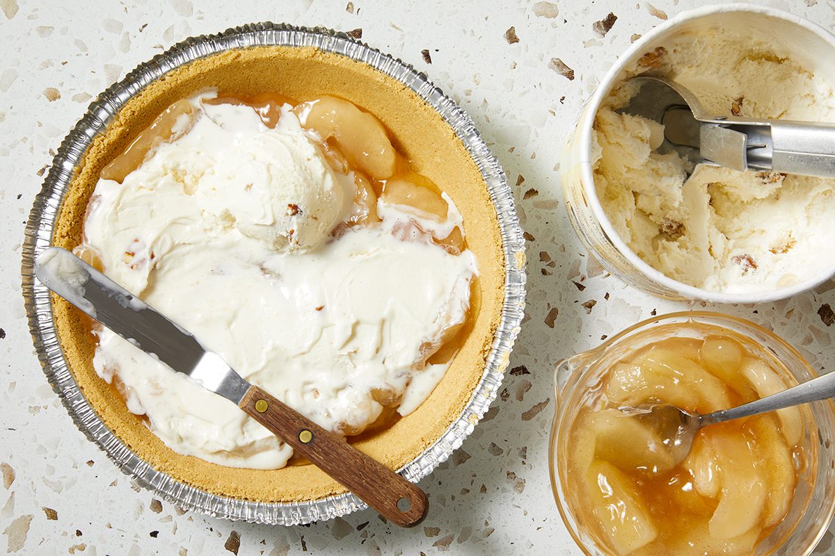 A graham cracker pie crust filled with vanilla ice cream and apple pie filling, next to a jar of apple filling and a tub of ice cream, all on a speckled countertop.
