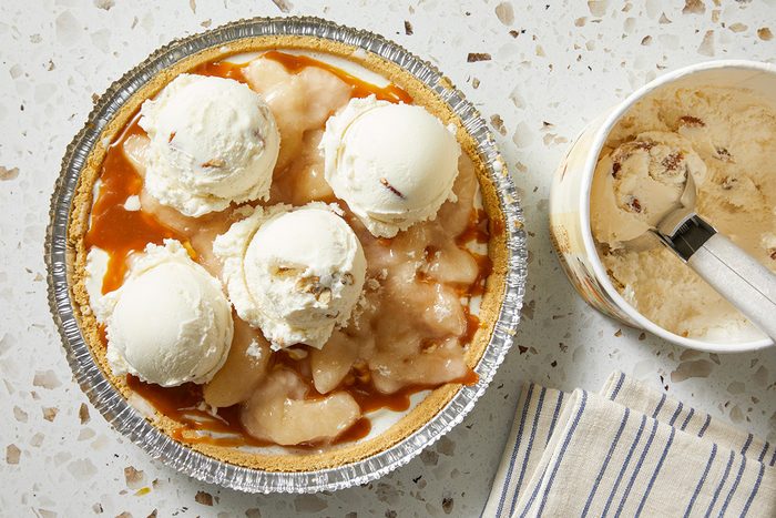 A pie in a foil pan topped with several scoops of vanilla ice cream and caramel sauce, next to a container of ice cream with a scoop inside, on a speckled countertop with a striped cloth nearby.