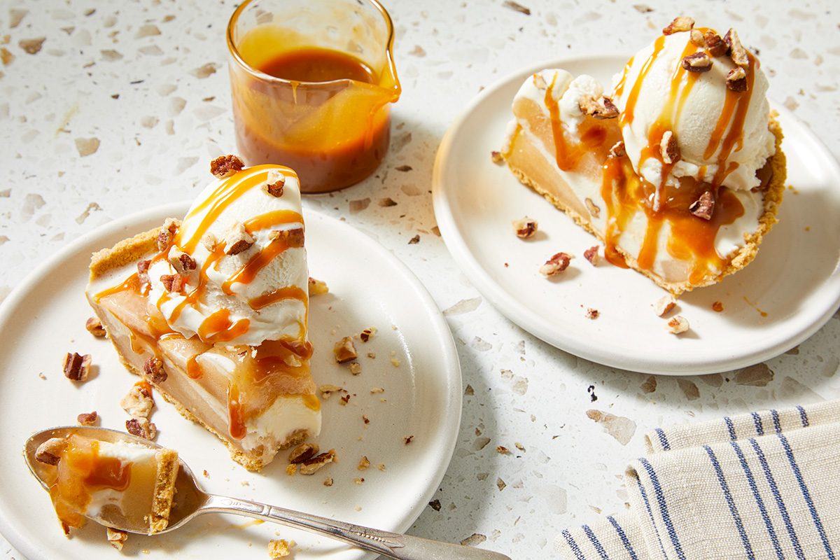 Two plates with slices of pie topped with vanilla ice cream, caramel sauce, and chopped pecans. A fork rests by one slice, and a small glass pitcher of caramel sauce is nearby on a terrazzo surface.