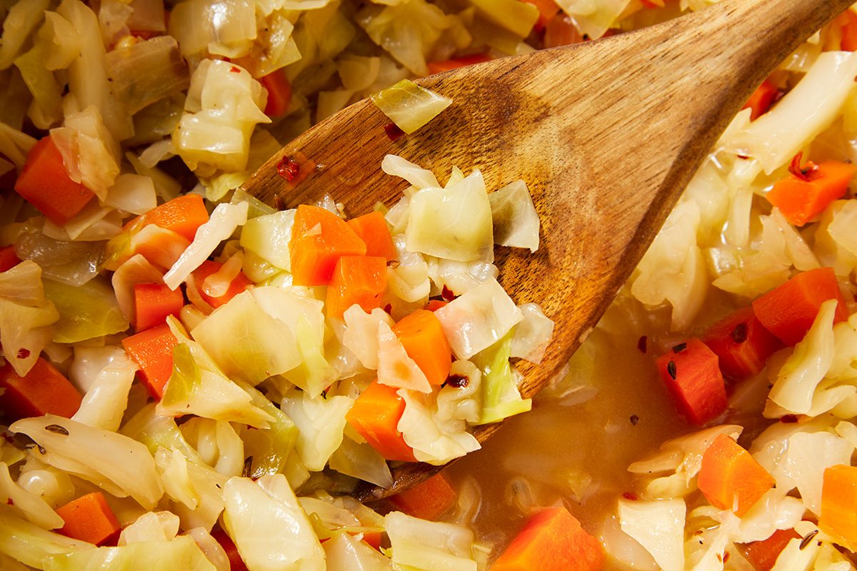 A close-up of cooked cabbage and carrots being stirred with a wooden spoon. The vegetables are chopped and appear to be seasoned, bathed in a light broth or sauce. The colors are vibrant, with orange carrots and pale green cabbage.
