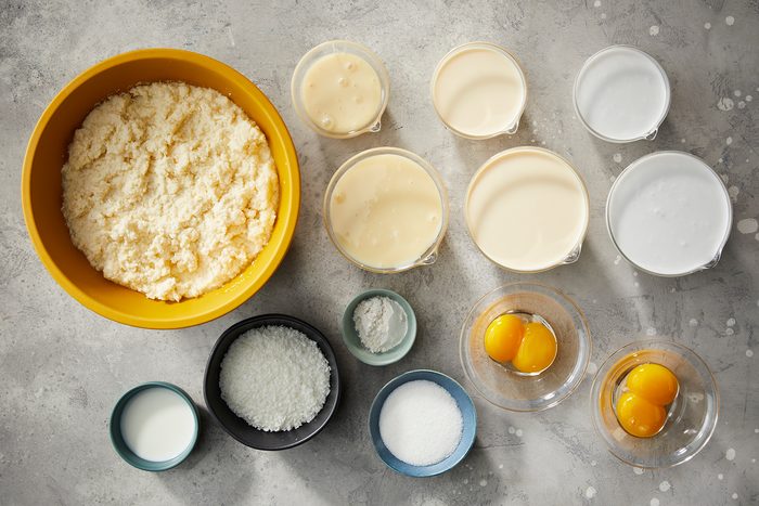 Various baking ingredients on a gray surface: a yellow bowl of cream, several clear bowls with liquid ingredients, small bowls containing shredded coconut, sugar, and flour, and two cracked eggs in separate glass bowls.