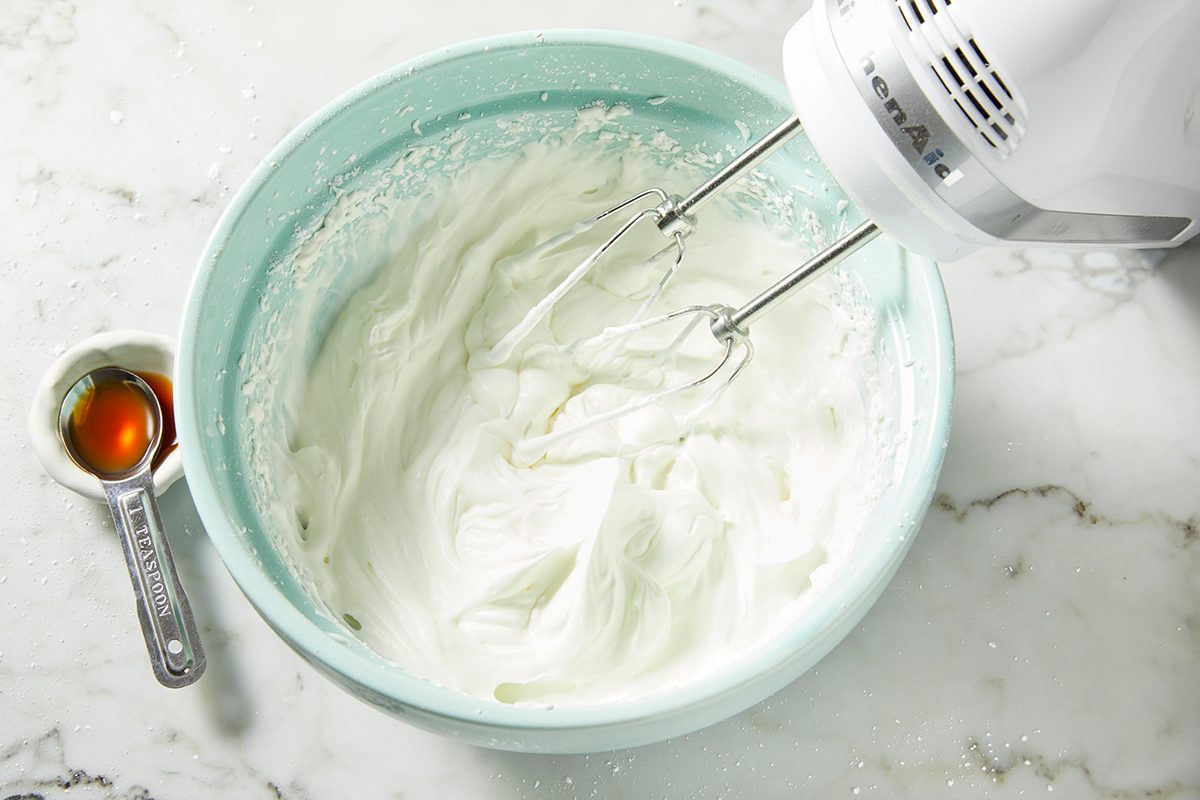 A white electric hand mixer whips fluffy cream in a light blue mixing bowl on a marble countertop. Beside the bowl, a measuring spoon filled with vanilla extract rests on the surface.