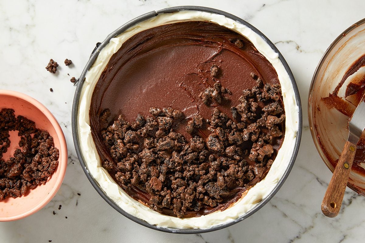 A dessert with a chocolate layer topped with crumbled cookies in a round pan, placed on a marble surface. Next to it are a bowl with additional cookie crumbs and a mixing bowl with a spatula.