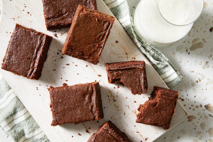 Overhead shot of Eggless Brownies; cut into pieces and served on white wooden tray over a napkin; A glass of milk is nearby; all set on a marble surface;