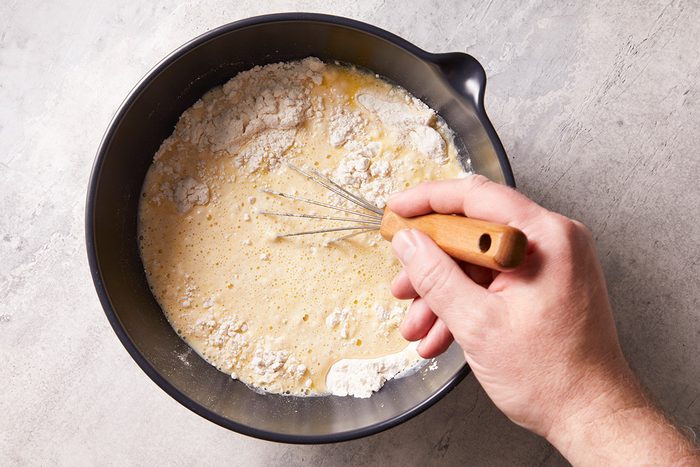 A hand holding a whisk mixes flour and liquid ingredients in a black bowl on a light countertop, preparing a batter or dough.