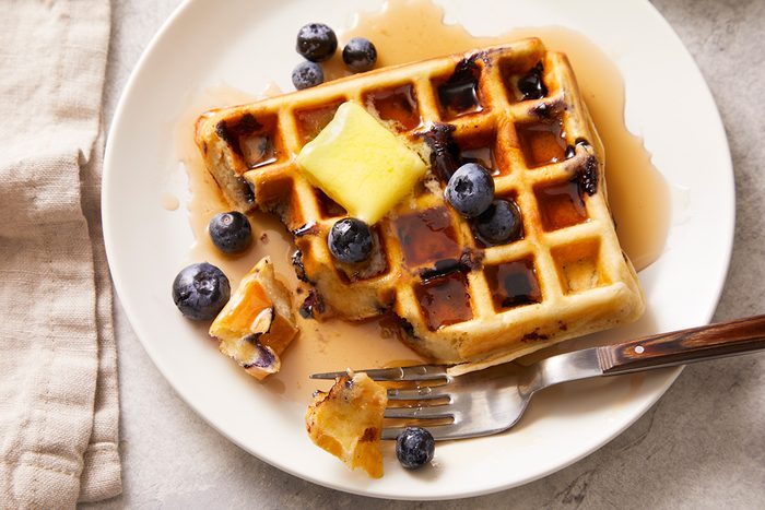 A partially eaten blueberry waffle topped with butter, syrup, and fresh blueberries sits on a white plate with a fork and beige napkin nearby.
