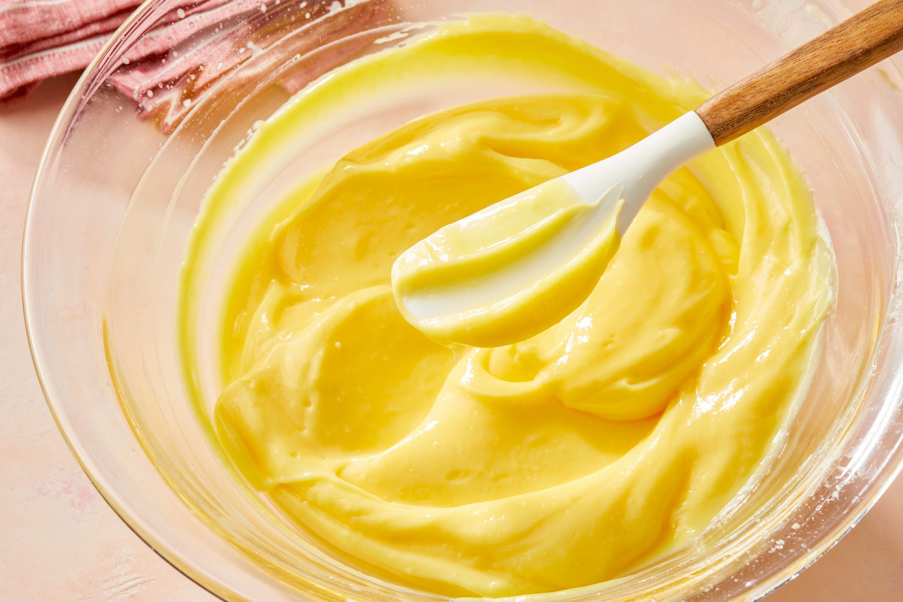 overhead shot of a clear glass mixing bowl filled butter, a white silicone spatula, with a wooden handle, is partially submerged in the mixture, in the background, there is a soft pink cloth visible