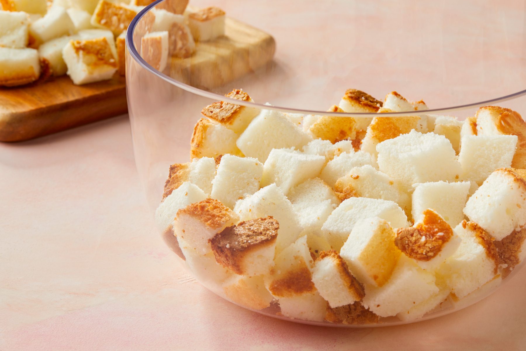 horizontal shot of a clear glass bowl filled with small, cubed pieces of bread; in the background, there is a wooden cutting board with additional bread cubes placed on it, the background has a soft, pinkish hue, surface