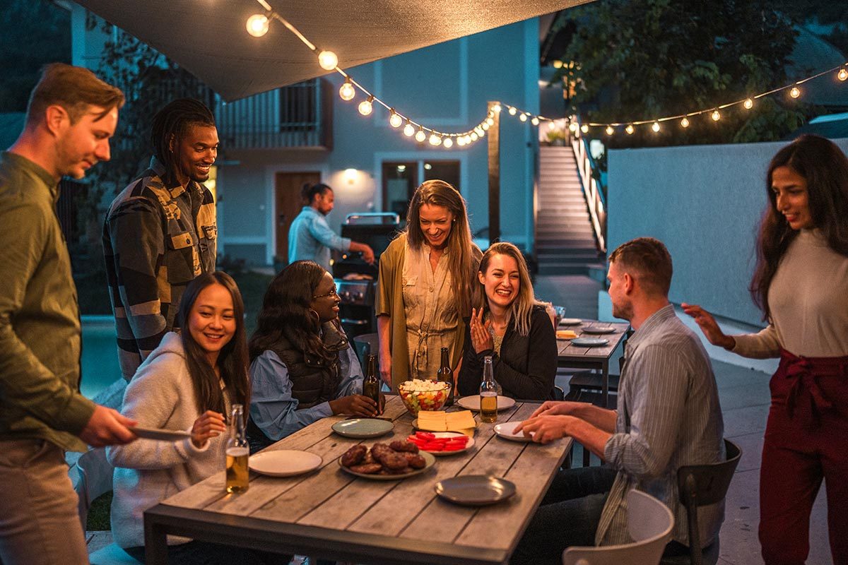 A diverse group of male and female friends gather around a table for an evening meal outdoors, enjoying drinks and conversation under string lights.
