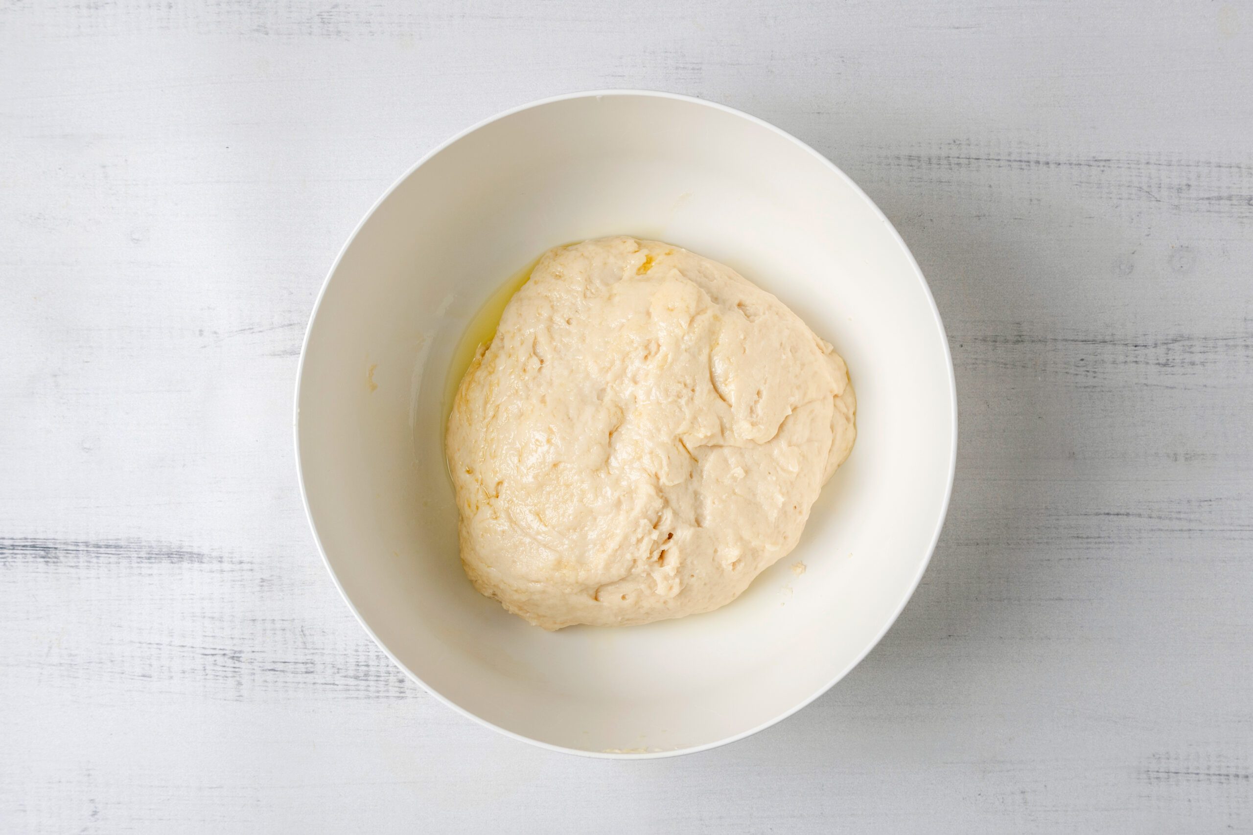 A ball of dough rests in a white bowl against a light gray background. The dough appears smooth and slightly glossy, indicating it's ready for further preparation or rising.