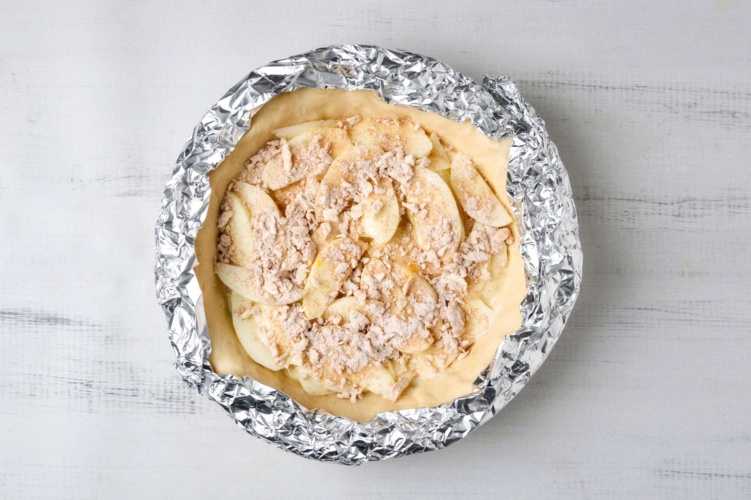 An unbaked pie with a crumb topping is wrapped in foil, sitting on a light gray surface. The pie contains visible apple slices and appears to be ready for baking.