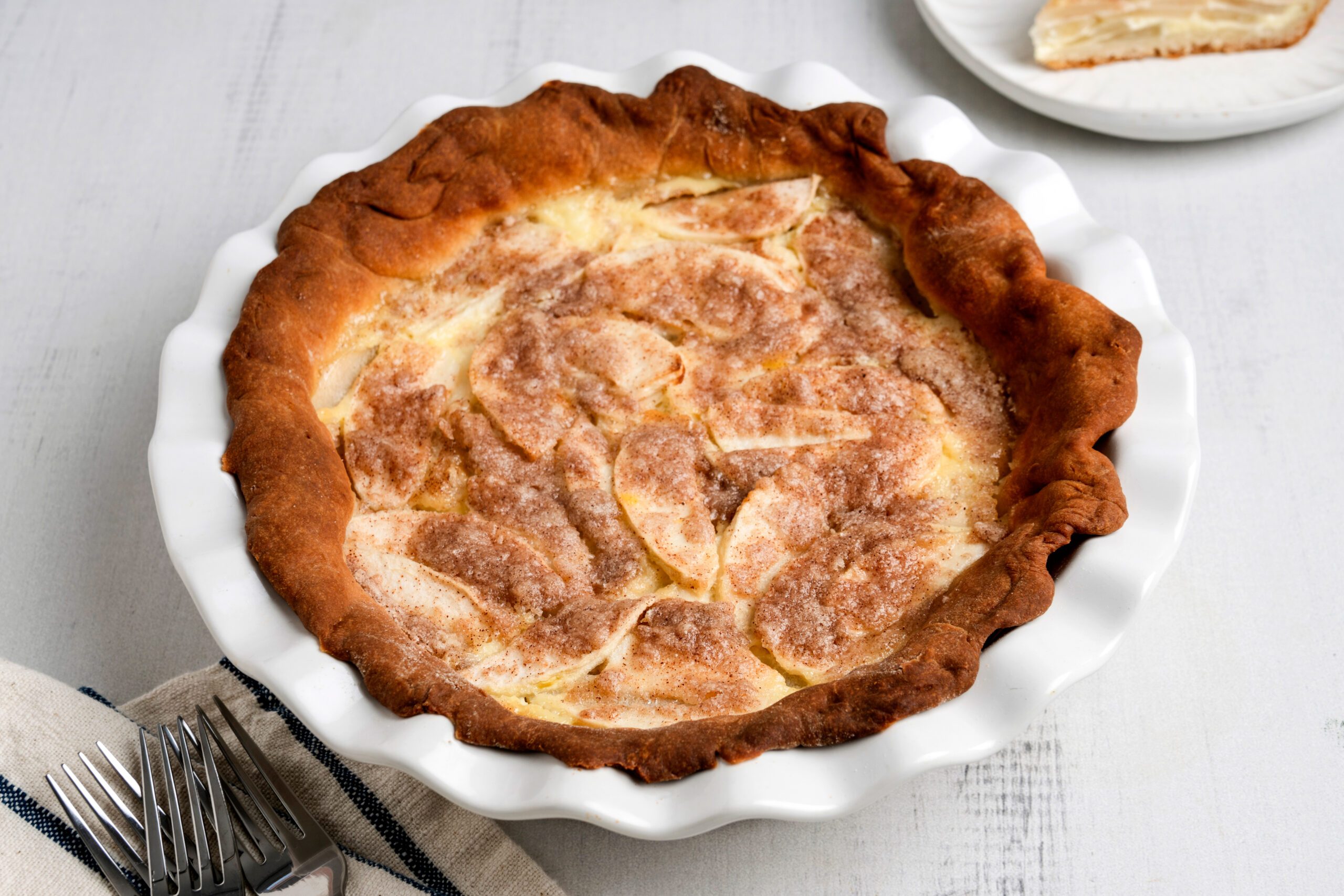 A freshly baked apple pie with a golden crust, sprinkled with sugar and cinnamon, sits in a white pie dish on a wooden table. 