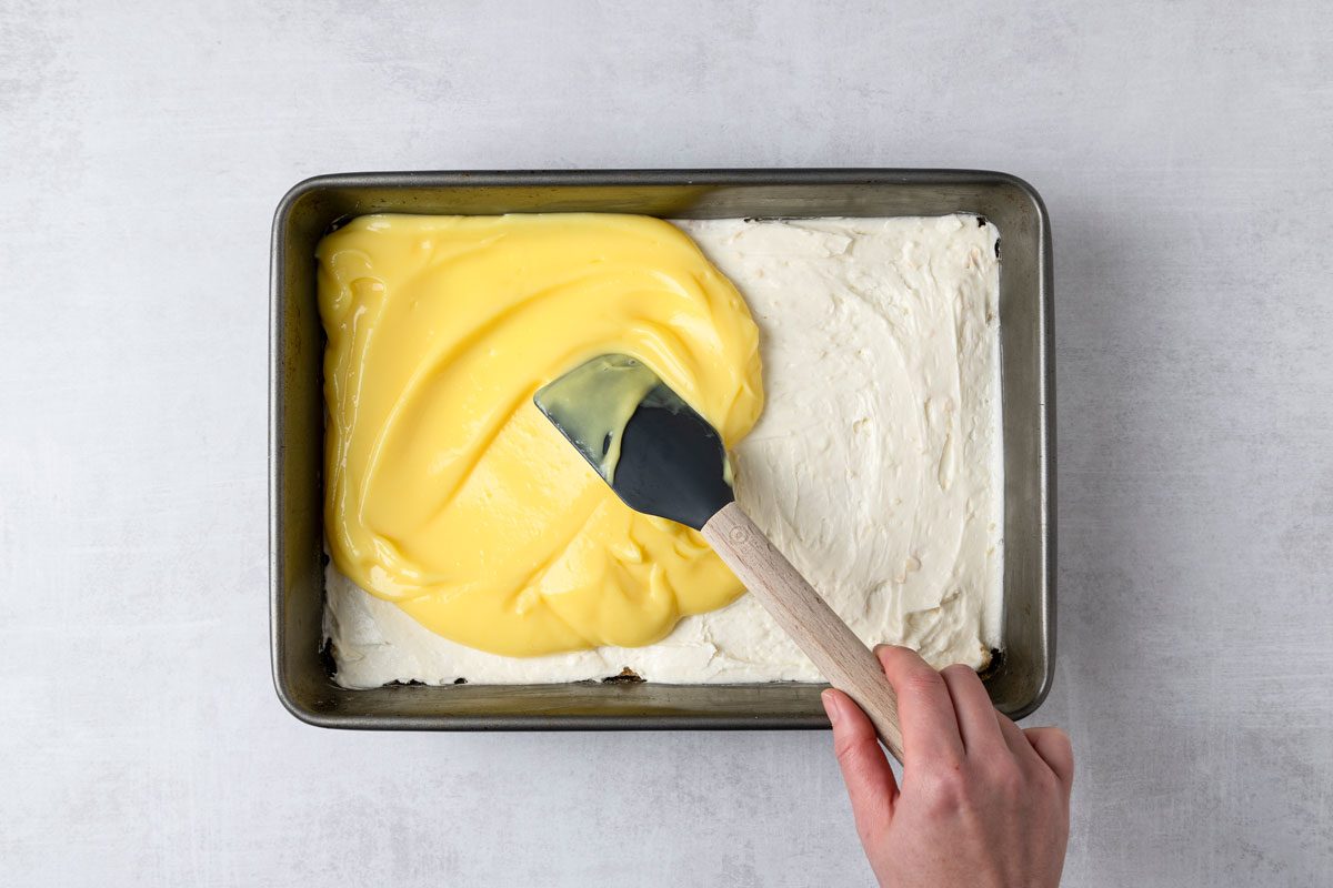 overhead shot of a baking pan with a layer of creamy white mixture on one side and a bright yellow custard filling being spread over the top on the other side; a hand holds a spatula, the background is a light grey