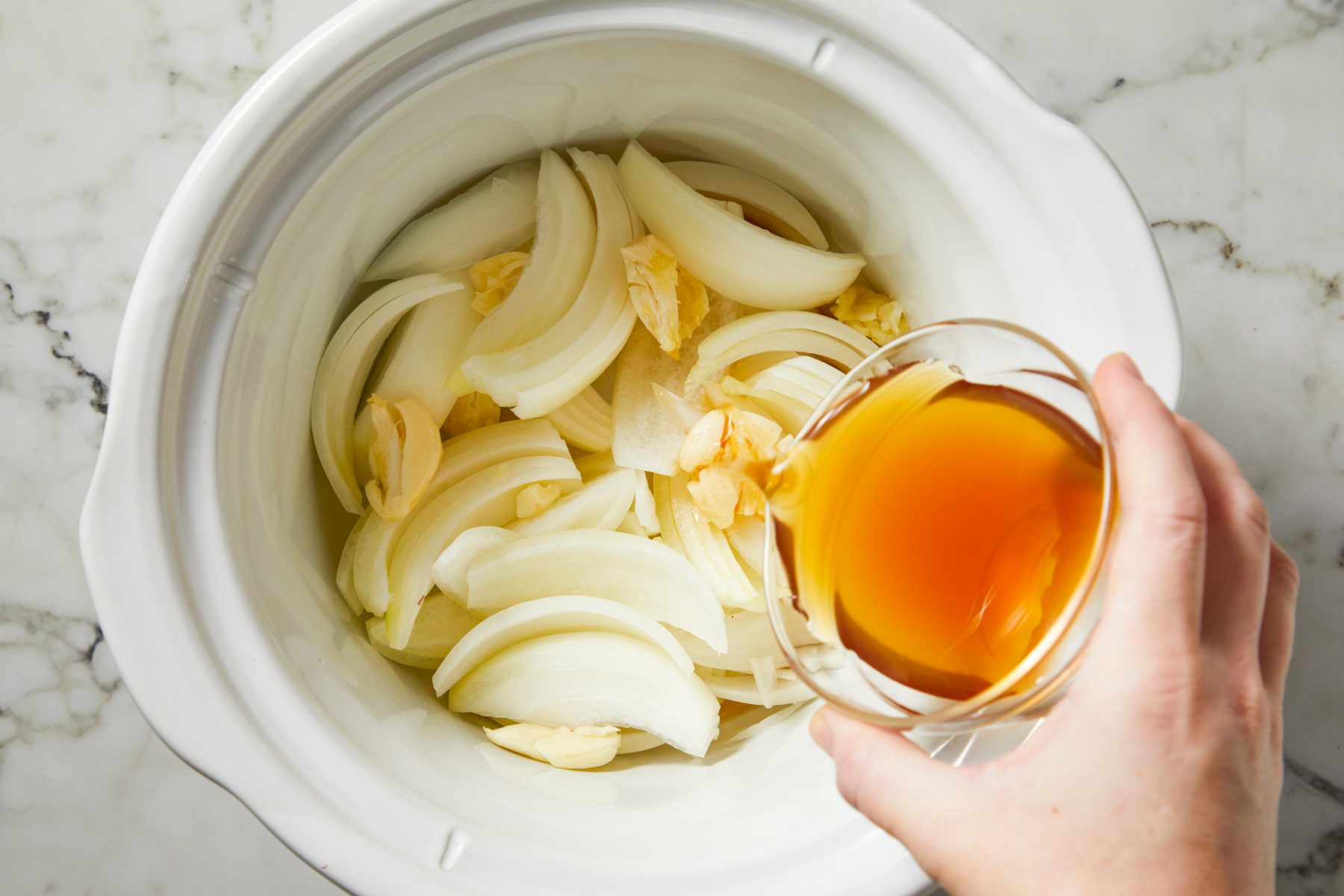 Onions and smashed garlic spread in slow cooker base, beef stock poured over.