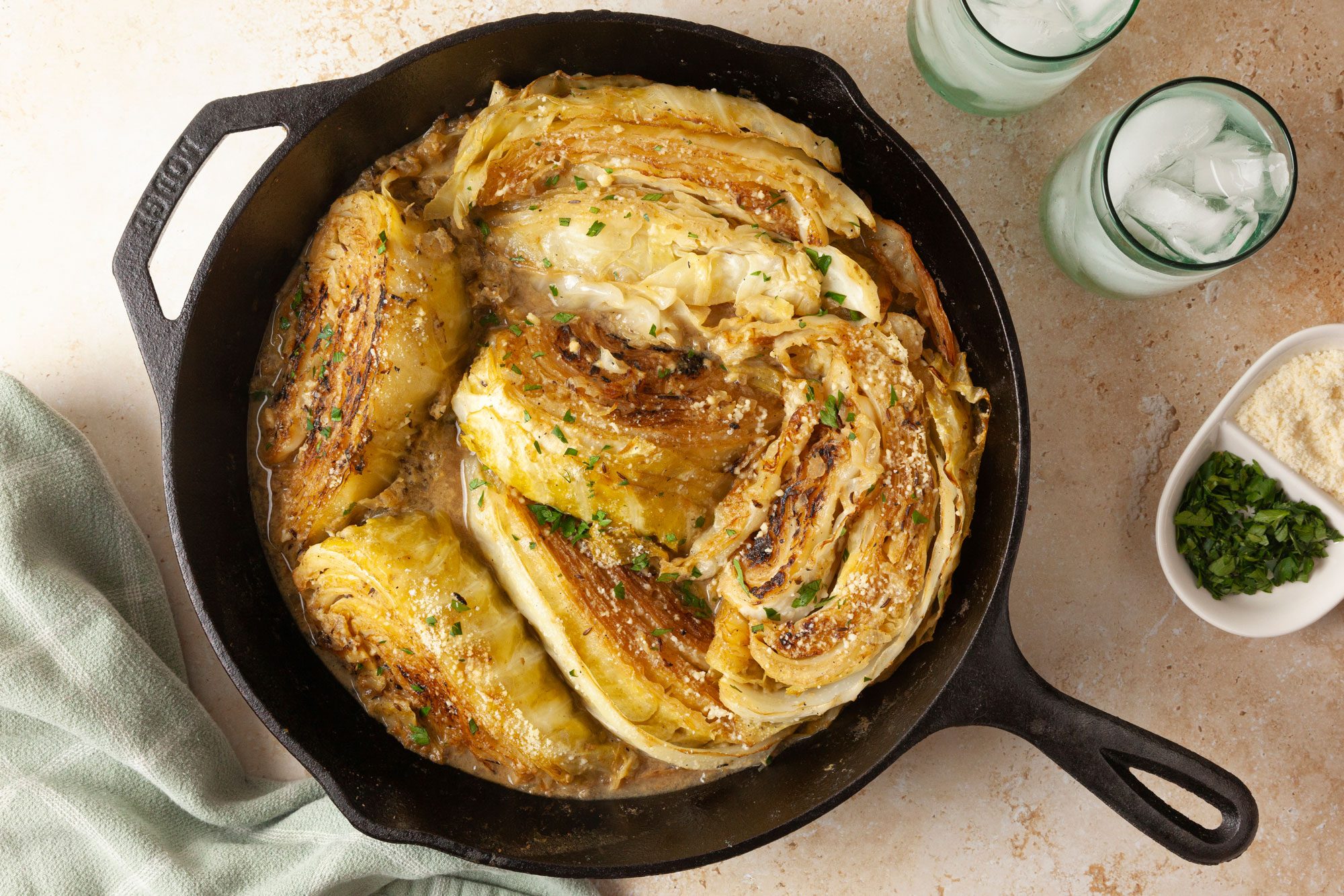 overhead shot of Melting Cabbage is presented in a cast iron skillet, beside the skillet, there are two glasses filled with ice and a soft green cloth is draped on the side; additionally, small bowls contain grated cheese and chopped herbs