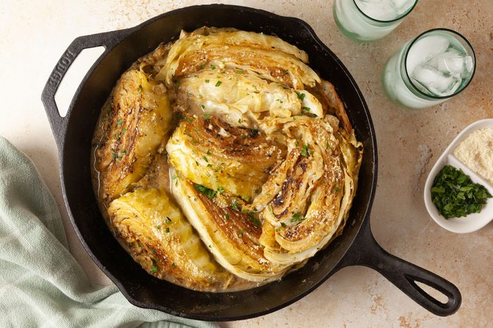 overhead shot of Melting Cabbage is presented in a cast iron skillet, beside the skillet, there are two glasses filled with ice and a soft green cloth is draped on the side; additionally, small bowls contain grated cheese and chopped herbs
