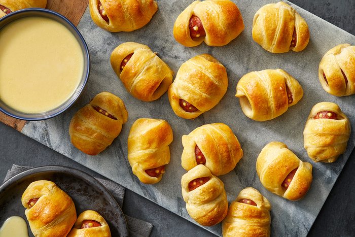 A marble board topped with several golden-brown pigs in a blanket, next to a bowl of yellow dipping sauce and a plate with more appetizers.