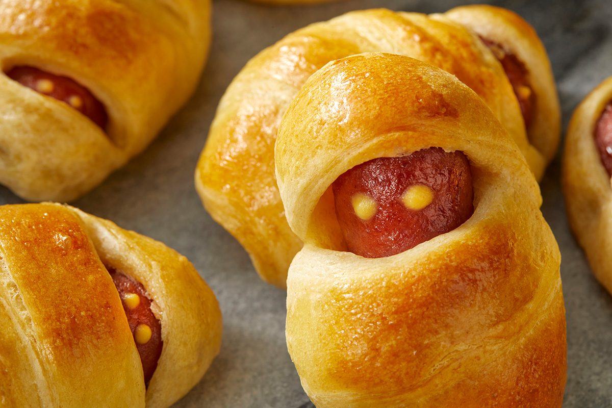 Golden-brown crescent rolls wrapped around mini sausages with small dots of mustard resembling eyes, arranged on a baking sheet.