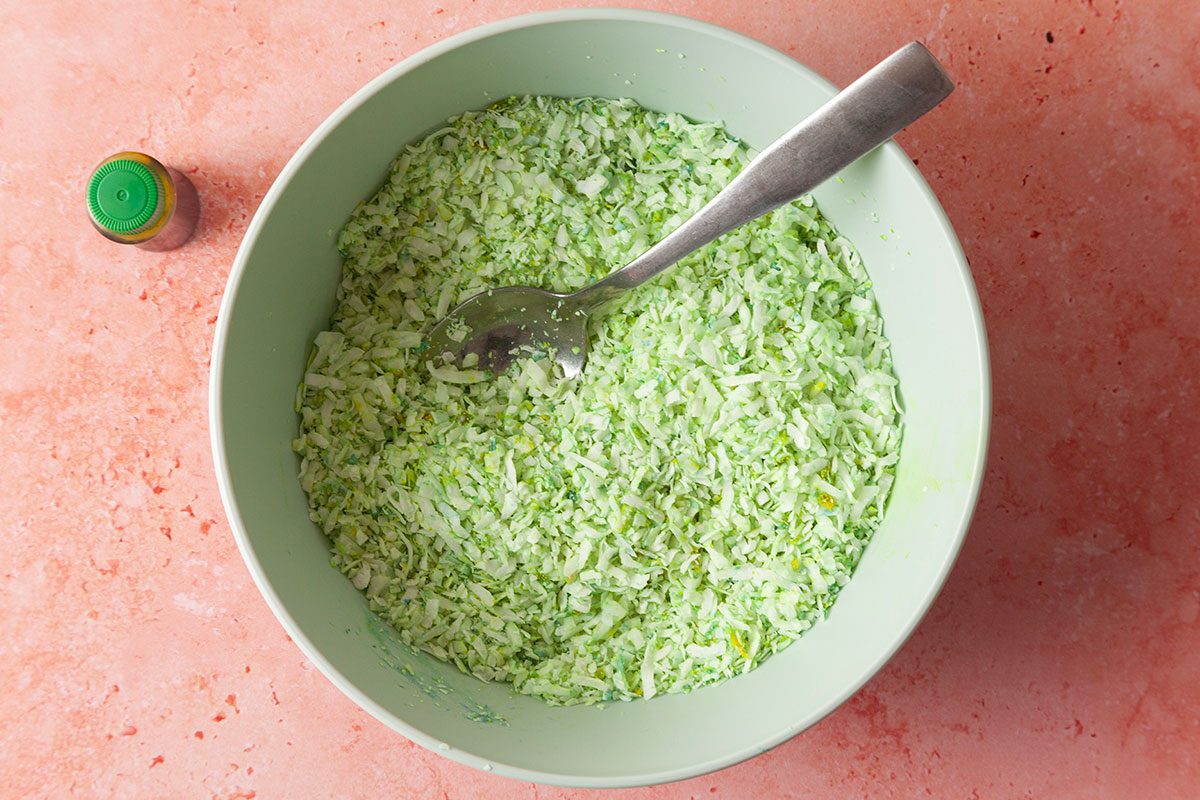 Bowl of green colored coconut flakes with a spoon in it next to a small vial of green food coloring