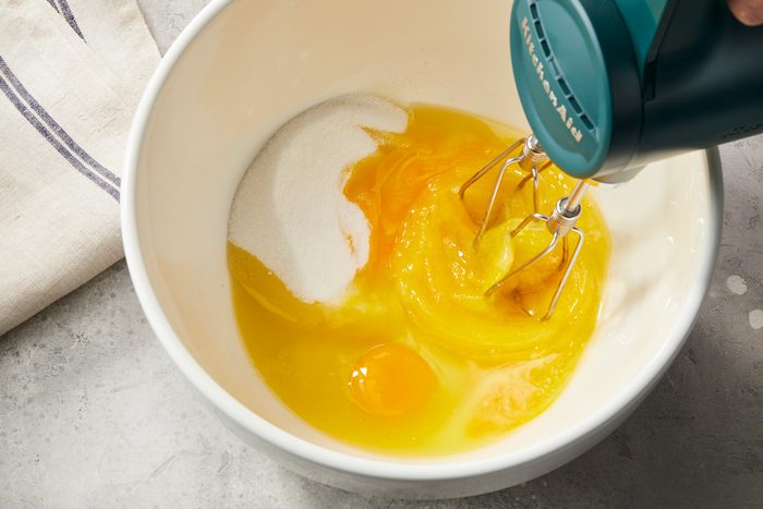 overhead shot of a white mixing bowl containing a mixture of eggs and sugar; several egg yolks are visible, along with one whole egg; a hand mixer is positioned above the bowl, with its whisks resting in the mixture; the setting appears to be a kitchen countertop