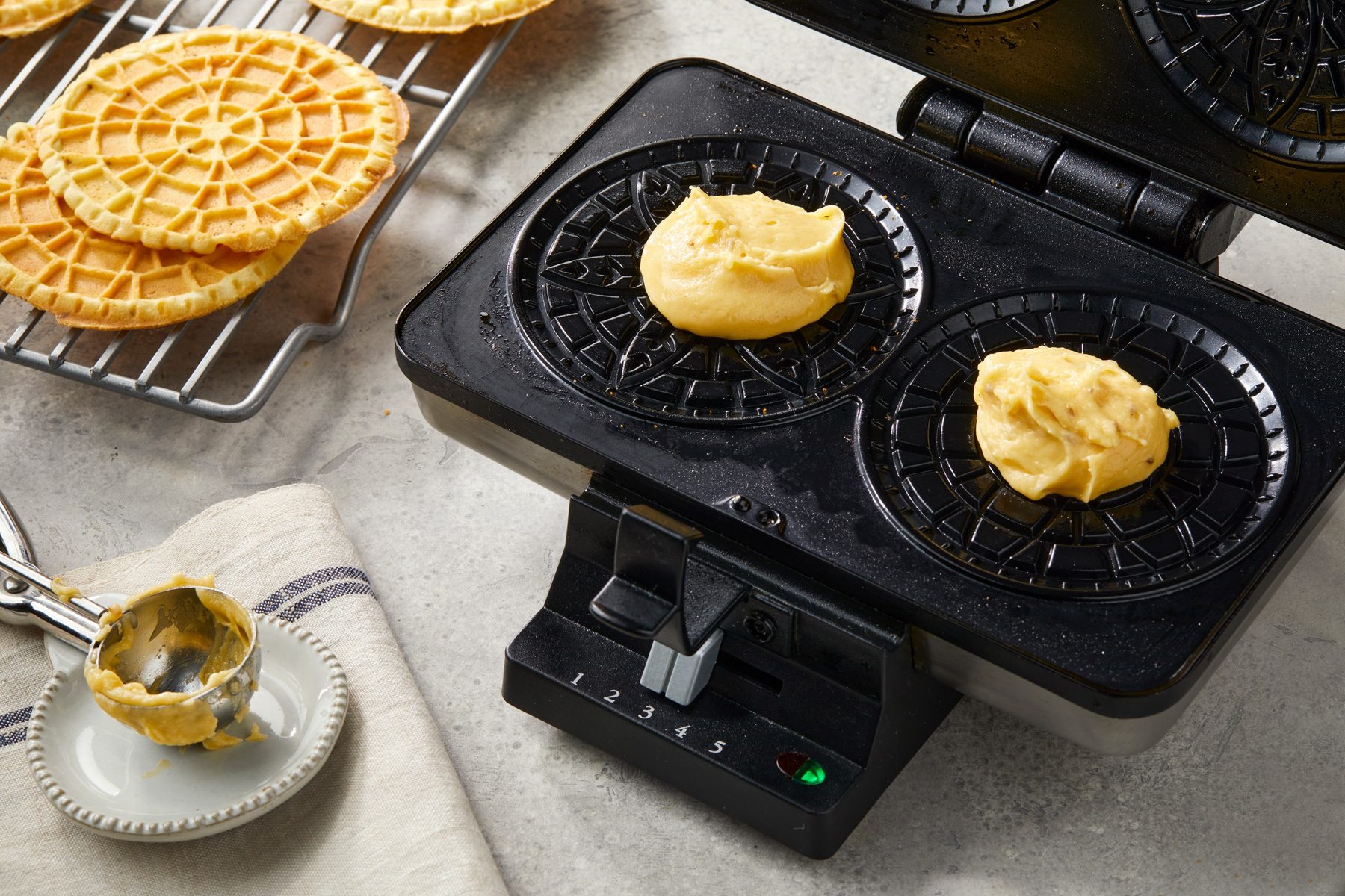 3/4th shot of a black waffle maker; inside, two small portions of batter are placed in the cooking chambers; next to the waffle maker, there is a cooling rack holding several golden brown waffles; a small spoon rests on a decorative plate, which contains a bit of the same batter; the background features a textured gray surface