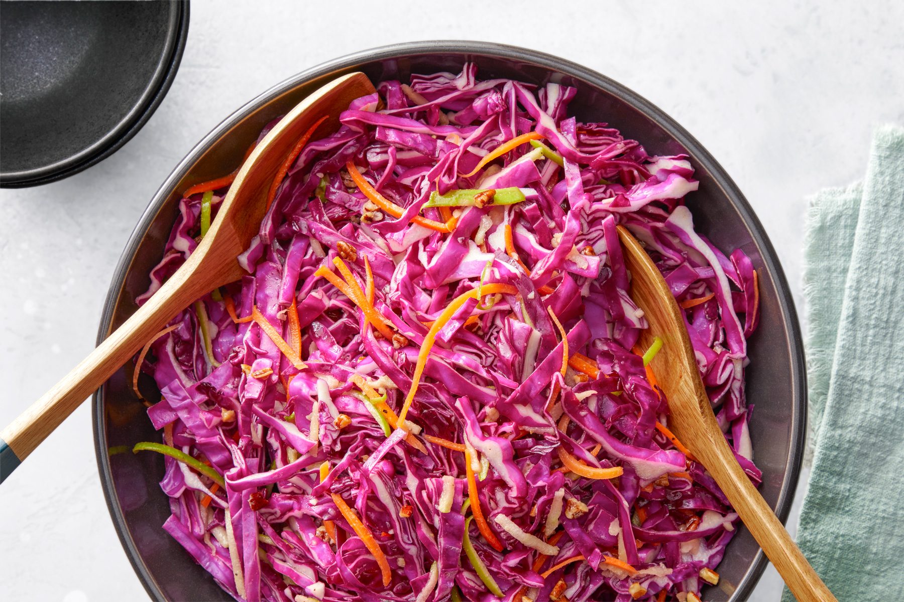overhead shot of Red Cabbage Salad in a large bowl, two wooden serving spoons are placed into dish, a light colored kitchen cloth is placed next to the main bowl; two dark colored bowls are slightly visible in background