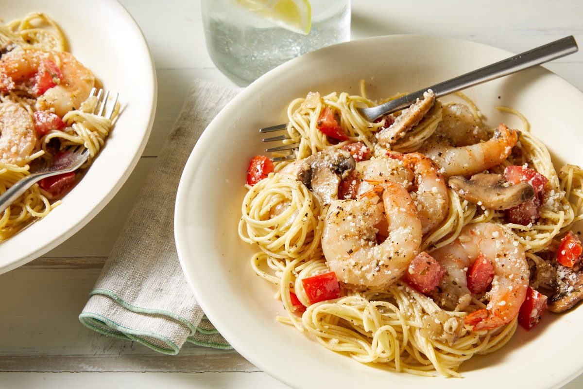 Close-up shot of Garlic Shrimp & Mushroom Pasta; served in two bowls with forks; sprinkle cheese on top; napkin; a glass of drink; white wooden surface;