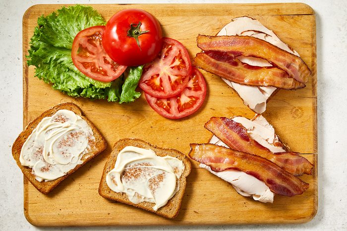 A wooden cutting board with slices of lettuce, tomato, turkey, bacon, and two pieces of bread spread with mayonnaise, ready to assemble into a sandwich.