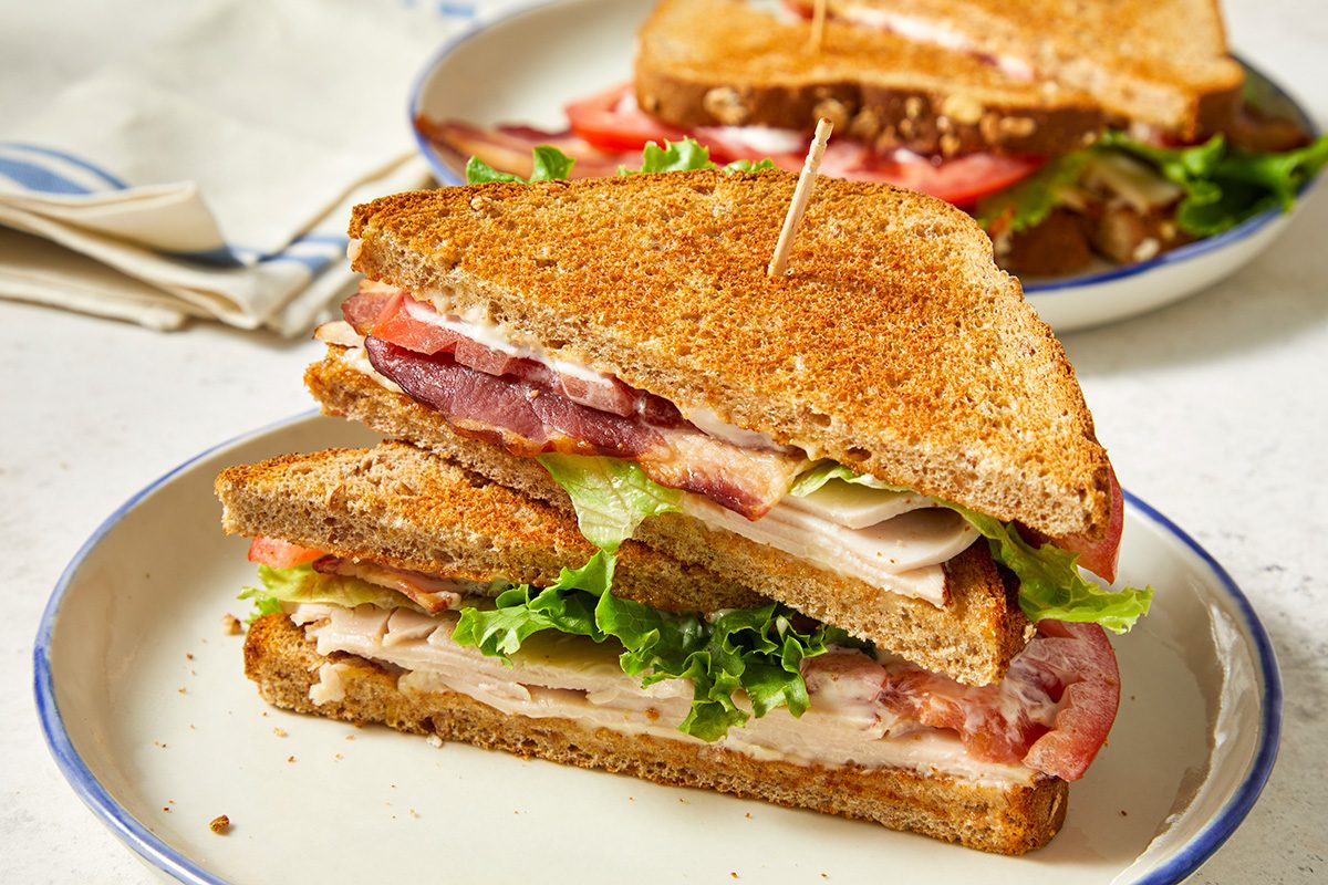A turkey sandwich with lettuce and tomato on toasted whole wheat bread, cut in half and stacked, served on a white plate with a toothpick holding it together. Another sandwich is blurred in the background.