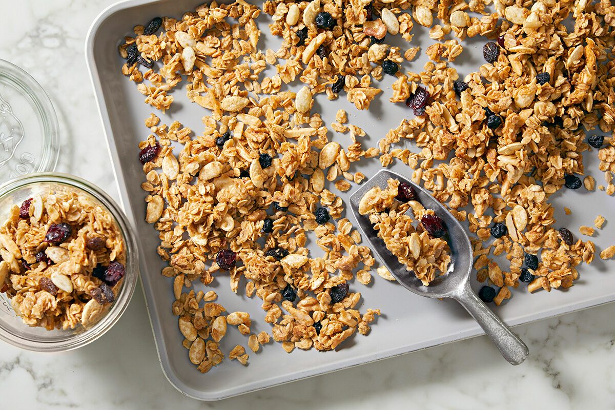 Granola spread out on a baking sheet with mixed nuts and berries, accompanied by a scoop and a jar filled with more granola. The baking sheet and jar are placed on a light marble surface.
