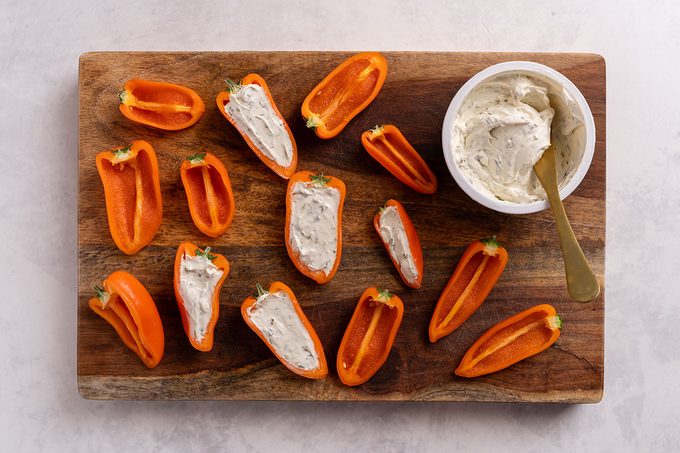 Orange bell peppers being filled with a cream cheese spread from a bowl, on a wooden cutting board