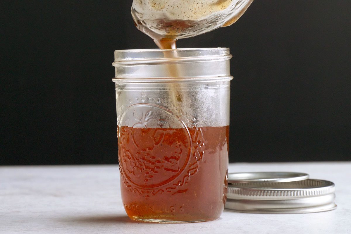horizontal shot of a mason jar is being filled with Apple Cinnamon Jelly that is being poured from a clear glass container; the mason jar is sitting on a white countertop, and the lid is sitting next to it;