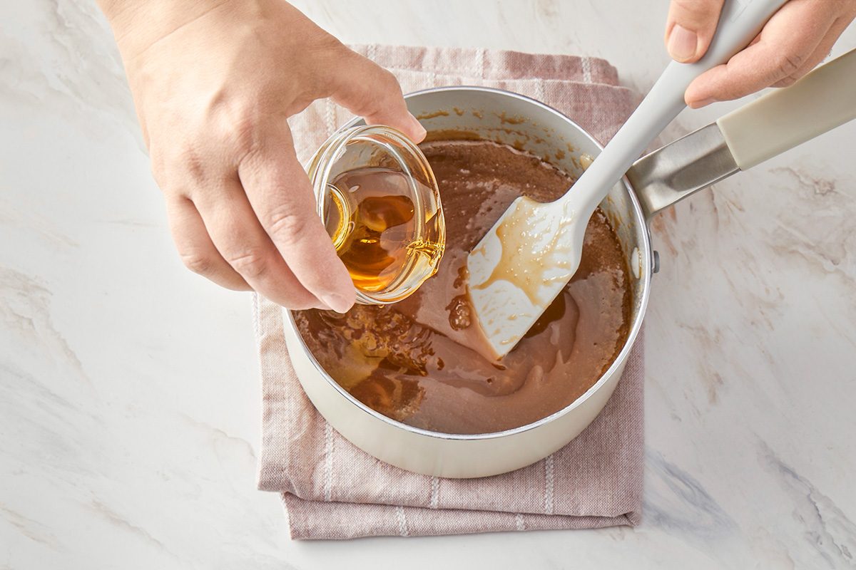 3/4th shot of A person pours a small glass bowl of vanilla extract, into a saucepan containing a light brown mixture, stirring with a spatula, The pan rests on a folded cloth on a marble surface