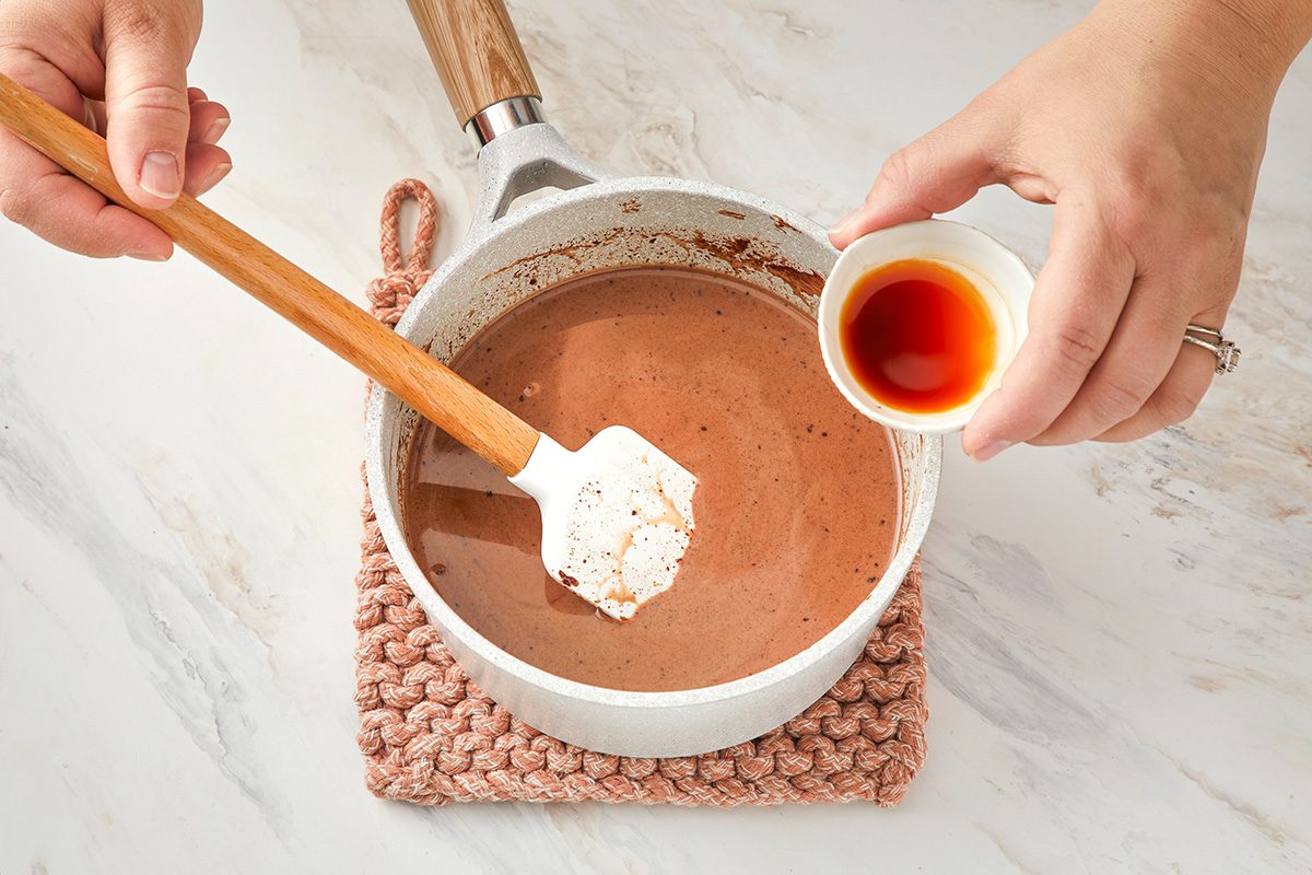 A person stirs a pot of chocolate mixture with a spatula while pouring vanilla extract from a small cup on a white countertop.