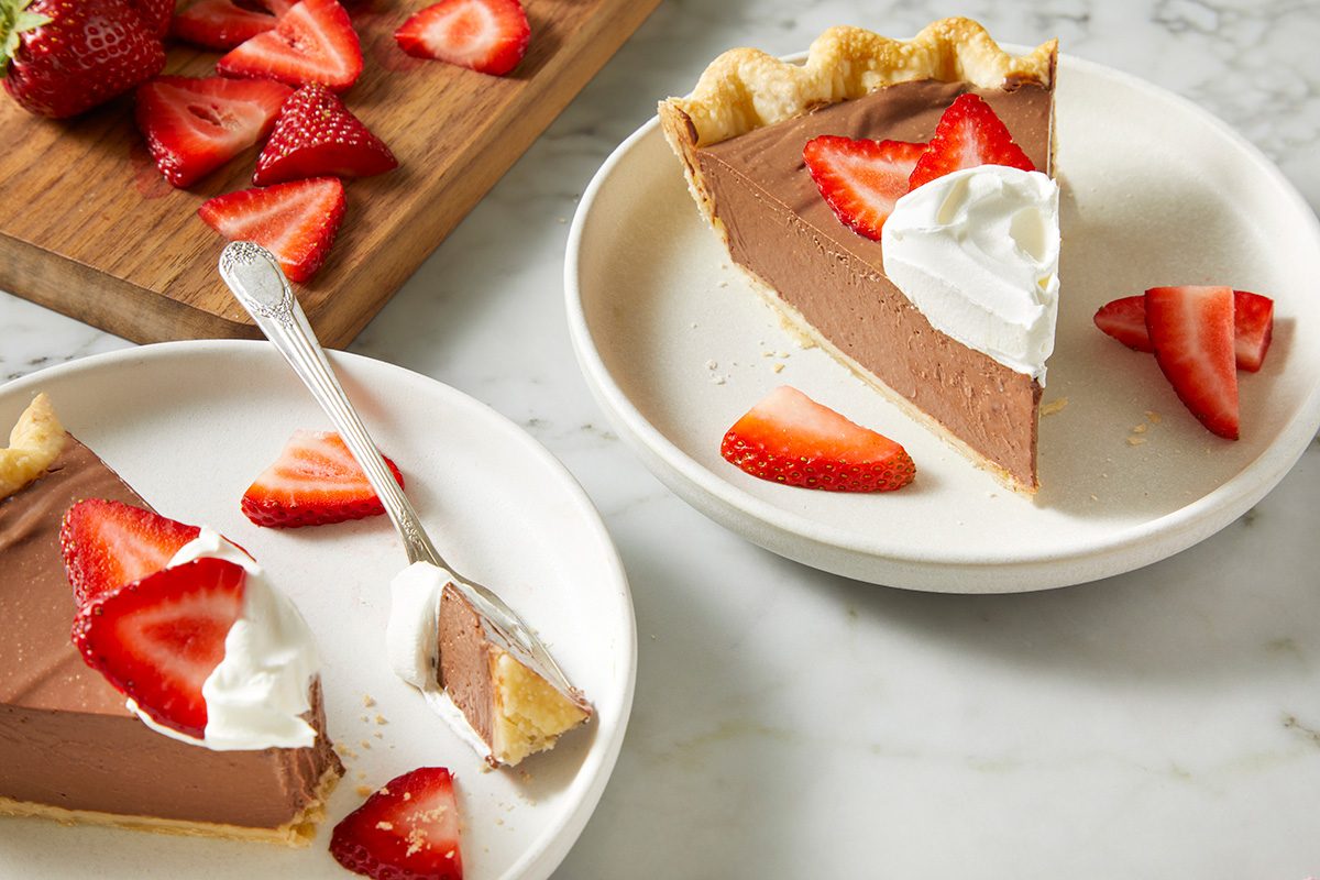 Two plates with slices of chocolate pie topped with whipped cream and fresh strawberry slices. A fork rests on one plate. A wooden board with sliced strawberries is in the background on a marble surface.