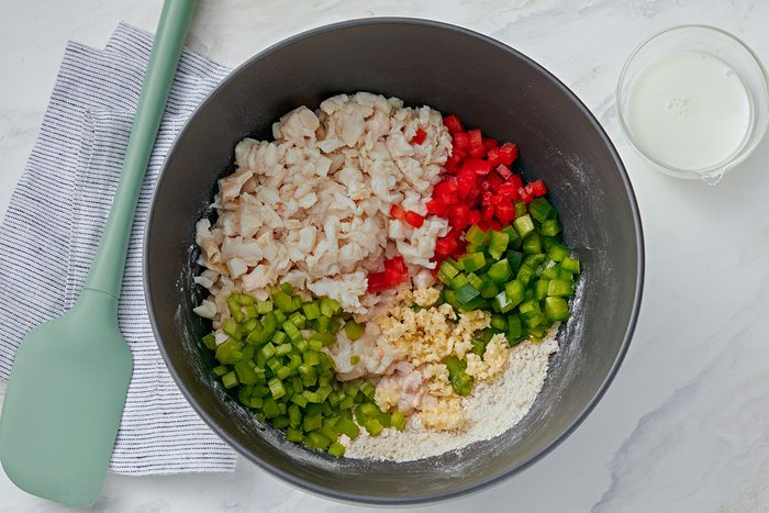 A mixing bowl with chopped shrimp, red and green bell peppers, celery, flour, and grated cheese, next to a green spatula, a striped cloth, and a small glass of milk on a white surface.