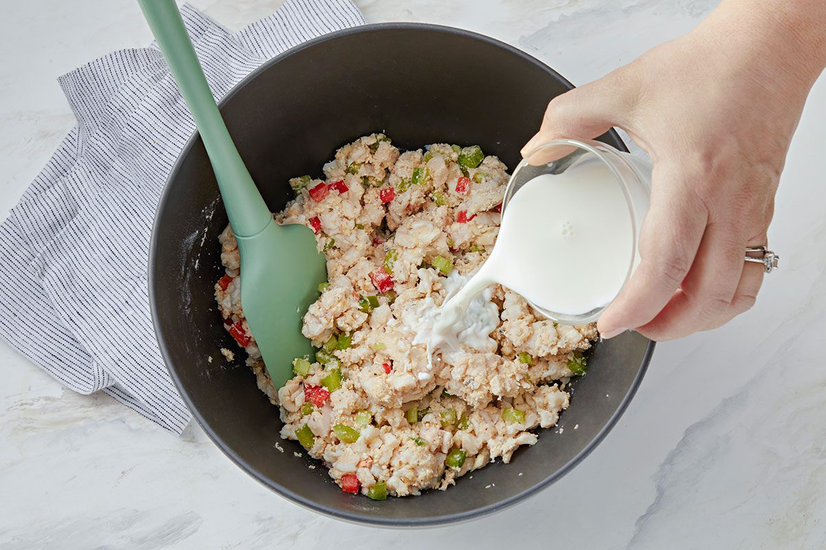 A hand pours milk from a small glass into a mixing bowl containing a mixture of rice, vegetables, and possibly crab, while a green spatula rests in the bowl. 