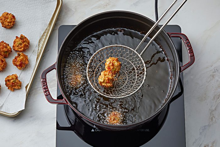 A wire skimmer holds a fried food ball above a pot of hot oil on a stovetop, with more fried balls cooking in the oil and several resting on a paper-lined tray to the side.