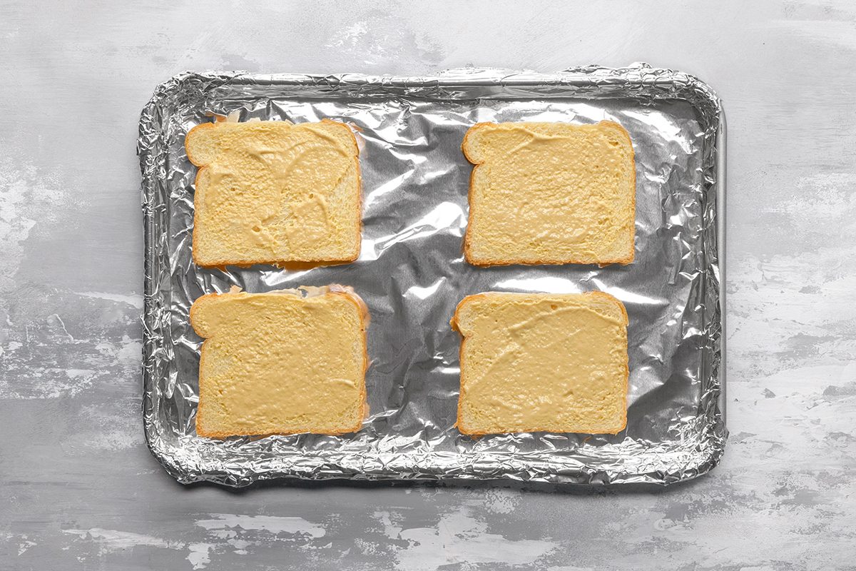 Four slices of bread spread with butter are placed on a foil-lined baking sheet, ready to be toasted.