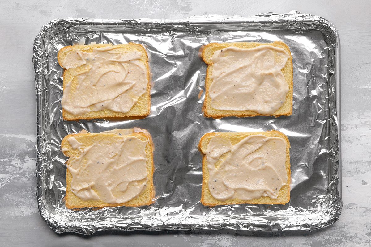 Four slices of bread topped with a creamy spread are arranged on a foil-lined baking sheet, ready to be baked.