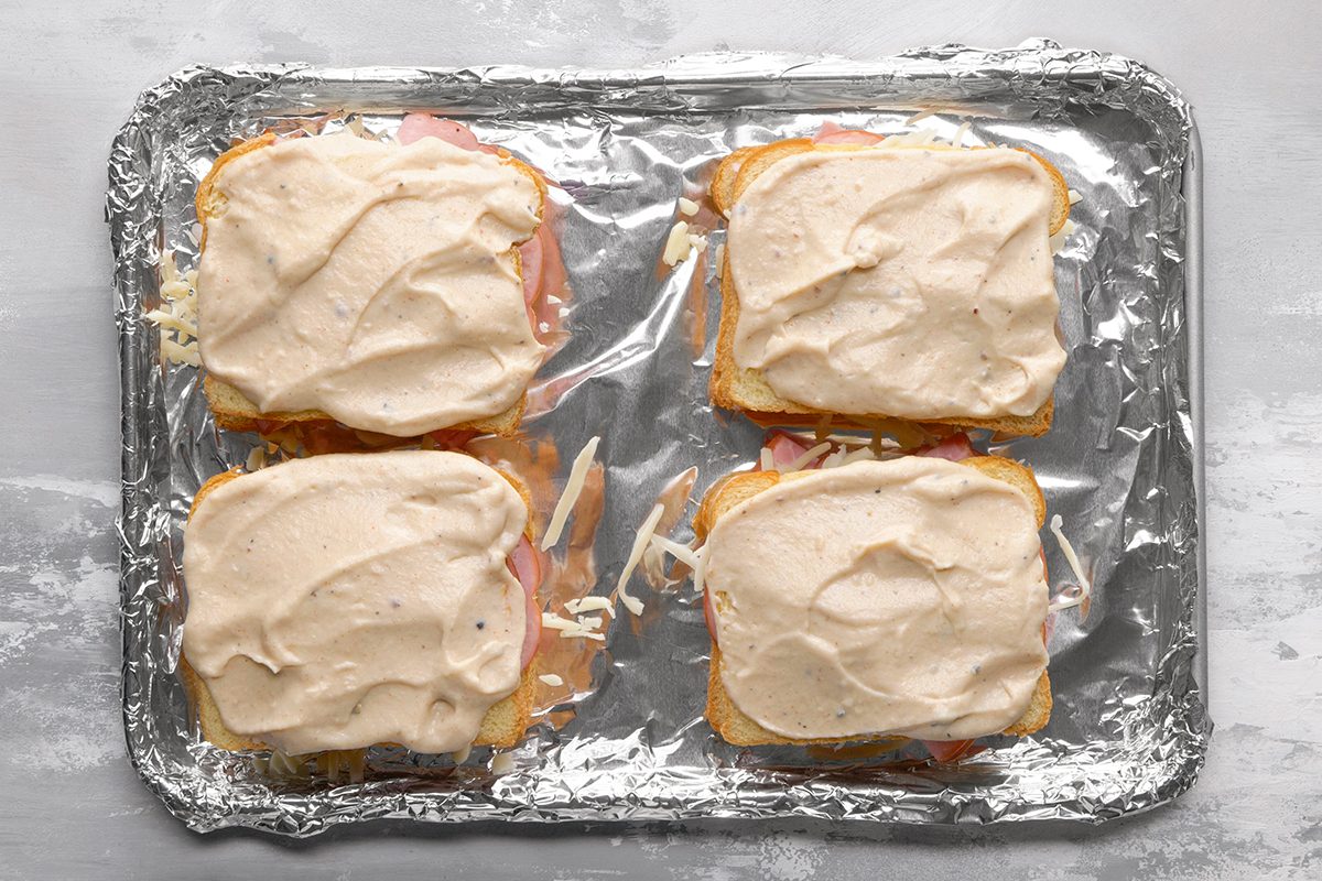 Four open-faced sandwiches topped with a creamy sauce are arranged on a foil-lined baking sheet, ready to be baked. The bread, filling, and sauce are visible, and the surface beneath the tray is light gray.