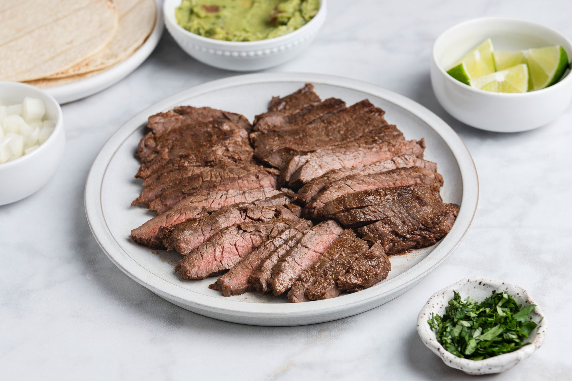 3/4th shot of Sliced grilled steak on a plate, surrounded by bowls of chopped onions, lime wedges, guacamole, chopped cilantro, and a stack of tortillas on a light marble surface
