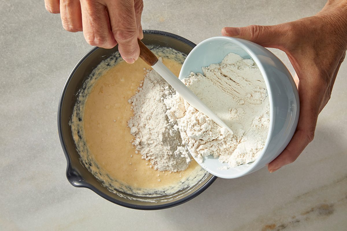 A person uses a spatula to mix flour from a blue bowl into a larger bowl containing a yellow batter on a light-colored countertop.