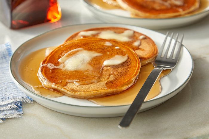 Two pancakes topped with melting butter and maple syrup sit on a white plate with a fork, set on a marble surface with a blue cloth napkin nearby.