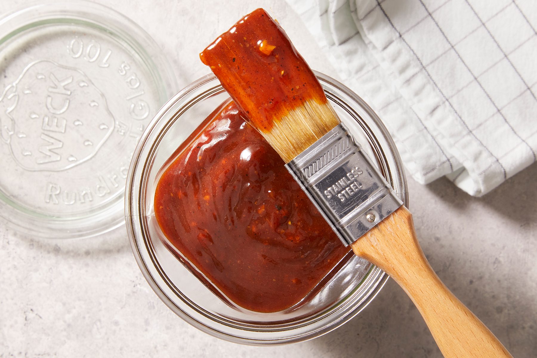 Top shot of barbeque sauce kept in a glass container with its lid beside it on top of a marble countertop with a check-pattern kitchen napkin kept beside it.