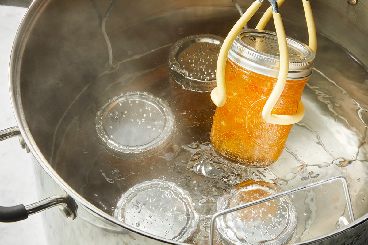 A canning jar of orange jam is being lifted with tongs from a pot of boiling water, surrounded by several sealed jars submerged in the water.