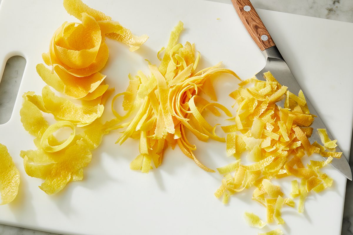A white cutting board with lemon peels arranged in three piles, from large strips on the left to finely chopped pieces on the right, and a knife with a wooden handle beside them.