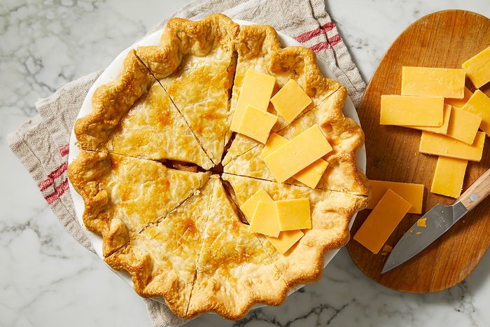 A baked pie with a golden, flaky crust sits on a towel. The pie is sliced, and several slices of cheddar cheese are placed on top and beside it, with a knife and more cheese on a wooden board nearby.